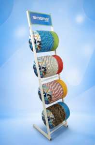 Fotografia com fundo branco de um Expositor Linha Tradicional da Riomar Cordas. É uma estrutura metálica vertical branca contendo 8 carretéis de cordas coloridas da Linha Tropical (nas cores verde, azul, amarelo, vermelho e padrões trançados). No topo, há um banner azul e verde com o logotipo da Riomar Cordas.