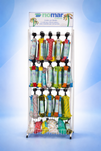 Fotografia Expositor Meada Riomar. Uma estrutura metálica vertical branca equipada com ganchos que exibem diversas meadas de cordas coloridas embaladas. No topo, há um banner com o logotipo da Riomar Cordas e a frase "Cordas em Meadas". Na base, o expositor possui um cesto de malha metálica para produtos adicionais.